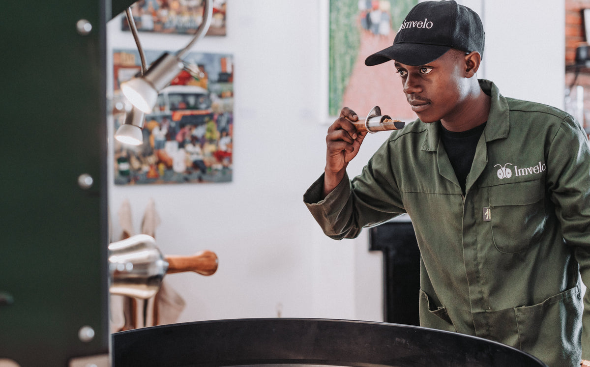 A roaster in Imvelo overalls and cap lifts a sample spoon from the drum and smells the beans at the roastery in Lusaka